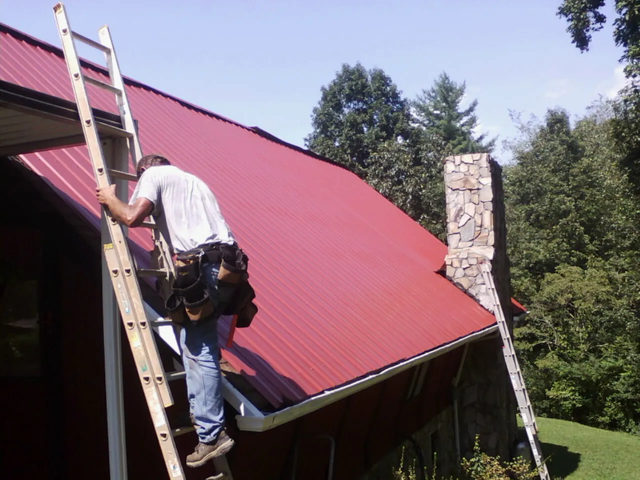 Roofer on a ladder beside a red metal roof and stone chimney — Soffit & Fascia Repair by Roofing Brothers in Fair Play